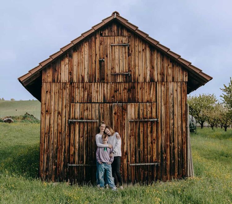 Drei Personen stehen vor einer Holzhütte im Gras und umarmen sich dabei.